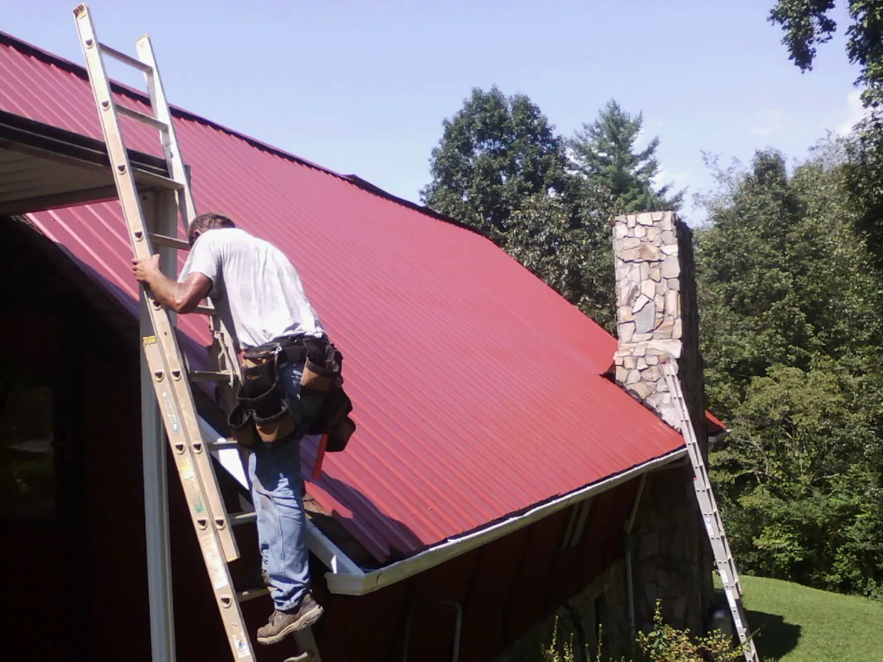 Roofer on a ladder beside a red metal roof and stone chimney — Soffit & Fascia Repair by Roofing Brothers in Mazon